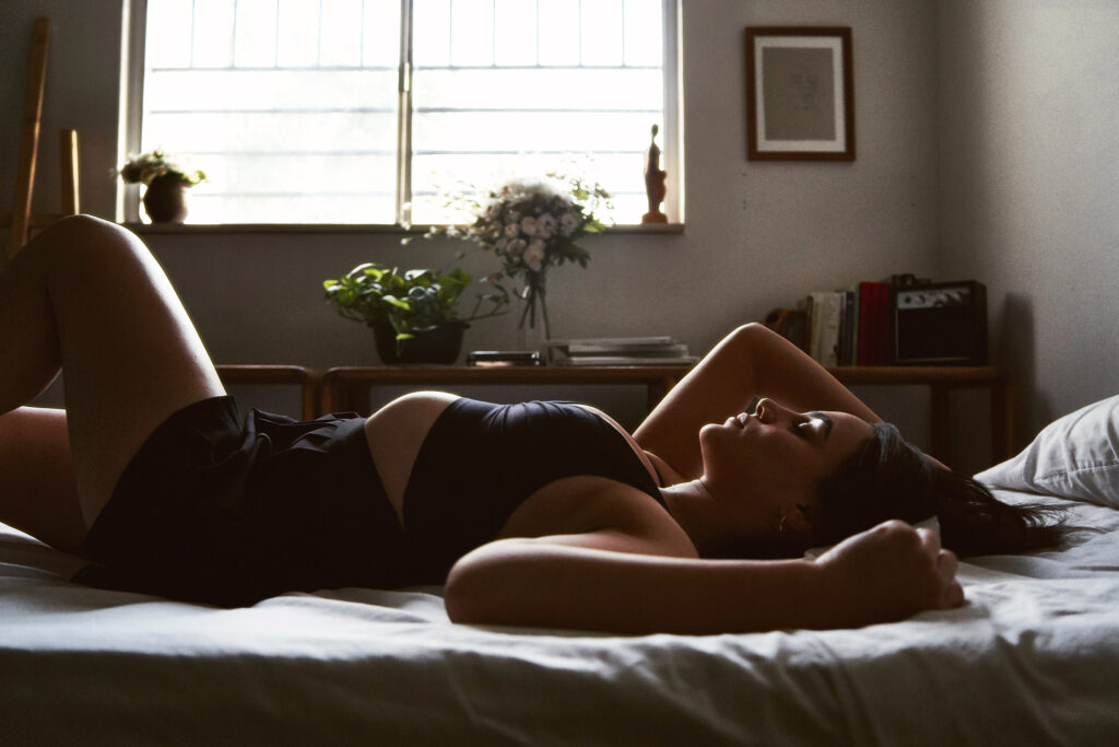 Valeria Cisneros lounges gracefully on a bed bathed in soft sunlight filtering through a window with a serene ambiance accentuated by indoor plants and personal items.
